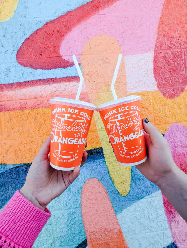 Two people cheersing with cups of Mackie's Orangeade in front of a brightly painted mural