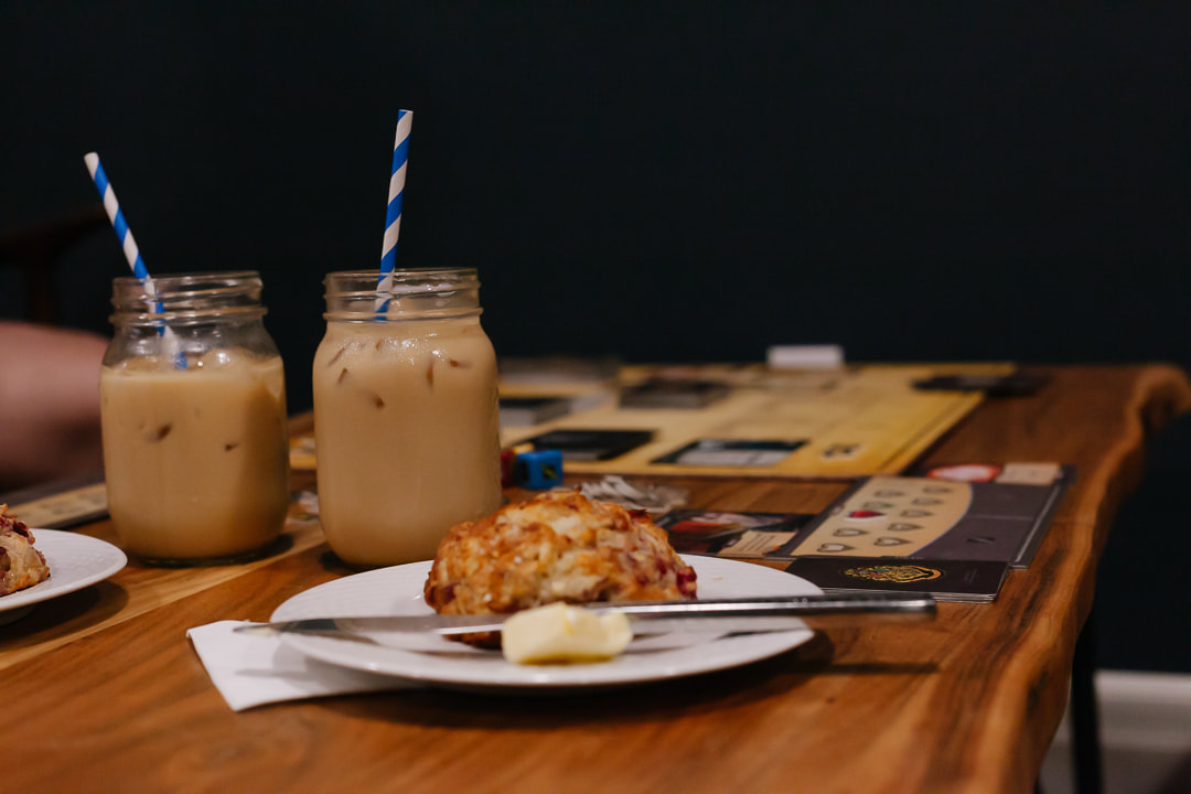 Iced coffee with blue and white paper straws in mason jars and a scone with butter sit on a table. There is a game also laid out on the table